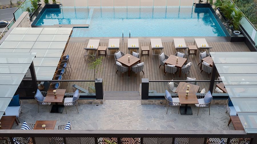 An aerial view shows a hotel rooftop with a swimming pool, lounge chairs, and several dining tables with chairs, arranged for guests to enjoy the outdoor space. - Iris Sarovar Portico, Sector 85, Gurugram