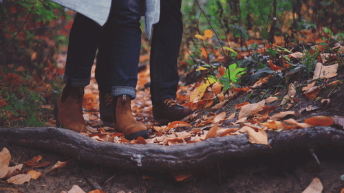 A close up shot of a couple wearing hiking boots in the woods with fallen dried leaves in view.