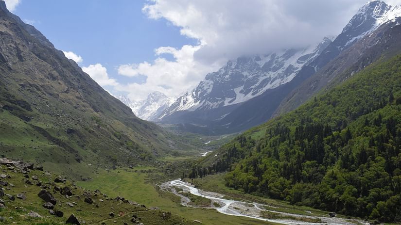 A breathtaking view of a wide mountain valley with a winding turquoise river flowing through it, framed by towering snow-dusted peaks under a vast sky with scattered clouds