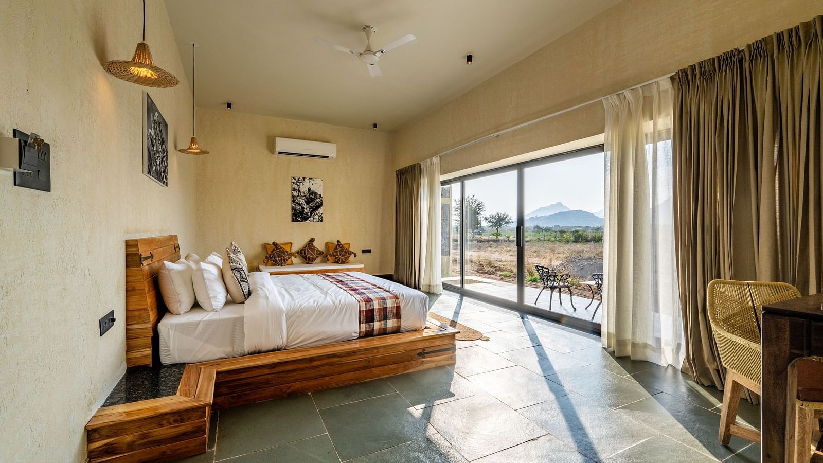  A spacious bedroom with a wooden study table with a chair, a bedside table, a wall-mounted air conditioner, photo frames, and a full-length window view at KURJA Jawai.