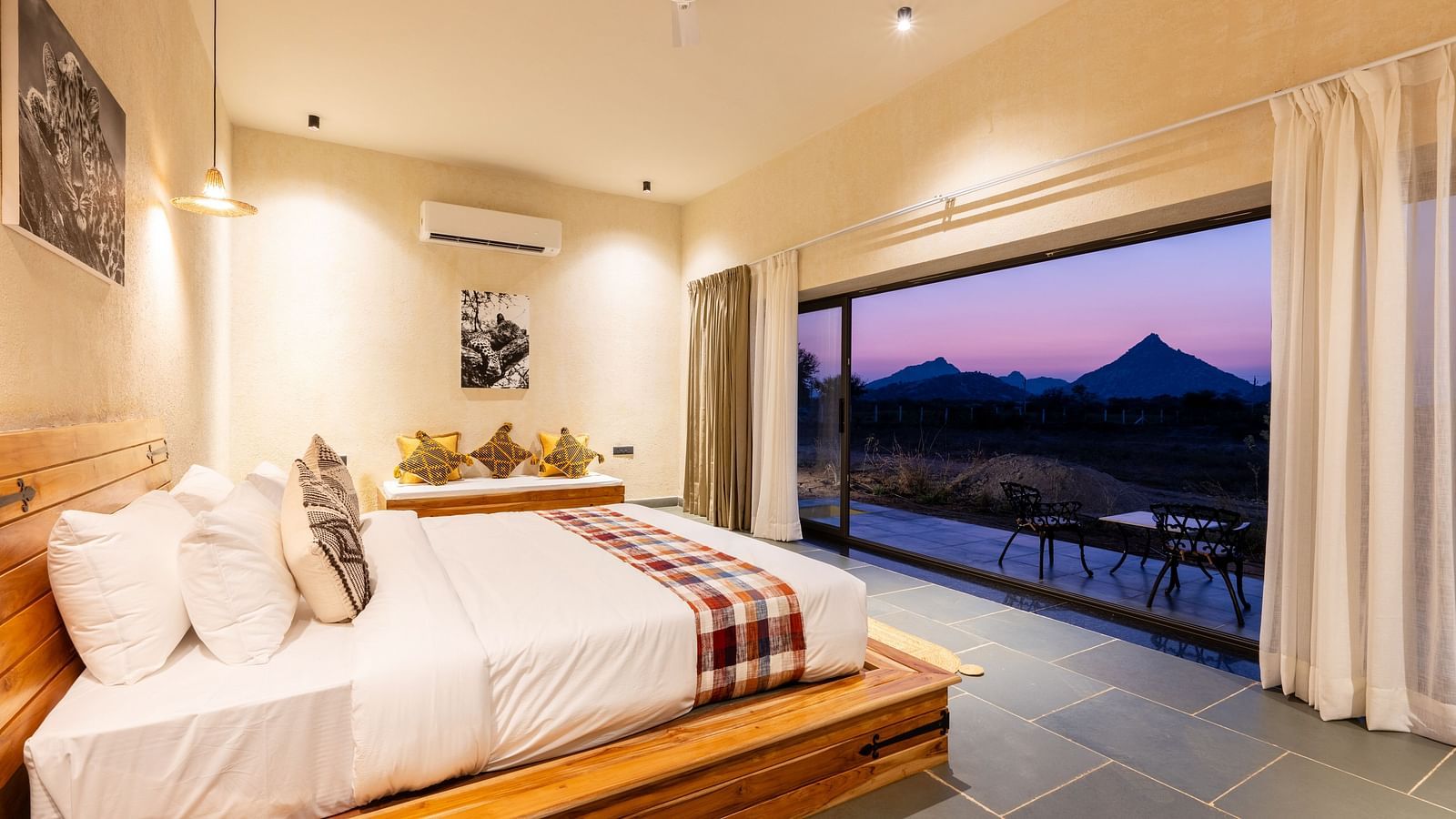  A spacious bedroom with a wooden study table with a chair, a bedside table, a wall-mounted air conditioner, photo frames, and a full-length window view  of mountains at KURJA Jawai.