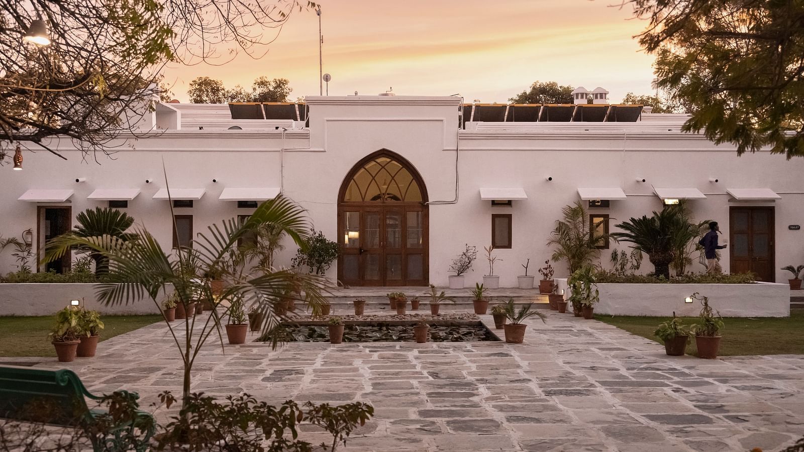 A white, single-story building with an arched doorway and paved courtyard under a warm sky at sunrise or sunset- Shahpura Bagh.