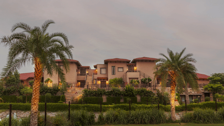 The resort's facade as seen from a distance at dusk, with lush greenery and palm trees under a cloudy sky at Aamaghati Wildlife Resort. Get in touch with Ranthambore Resorts Contact mentioning on the contact us page.