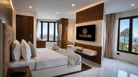 A Junior Suite AC Room at Valmere Chery Blossom Suites with a neatly-made bed, a wall-mounted television, wood-panelled walls, and big windows with full-length curtains.