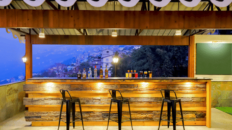 Bar at Valmere Himalayan Hotel & Spa, Gangtok with a wooden counter, bottles of spirits, 4 colourful cocktails, 3 stools, and a hillside town view at dusk.