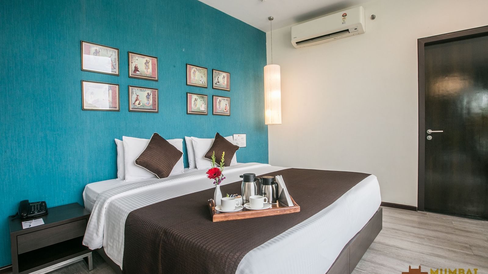 Hotel room with a turquoise feature wall, framed artwork, white bedding, and a brown bed runner, plus a tea service set on the bed.