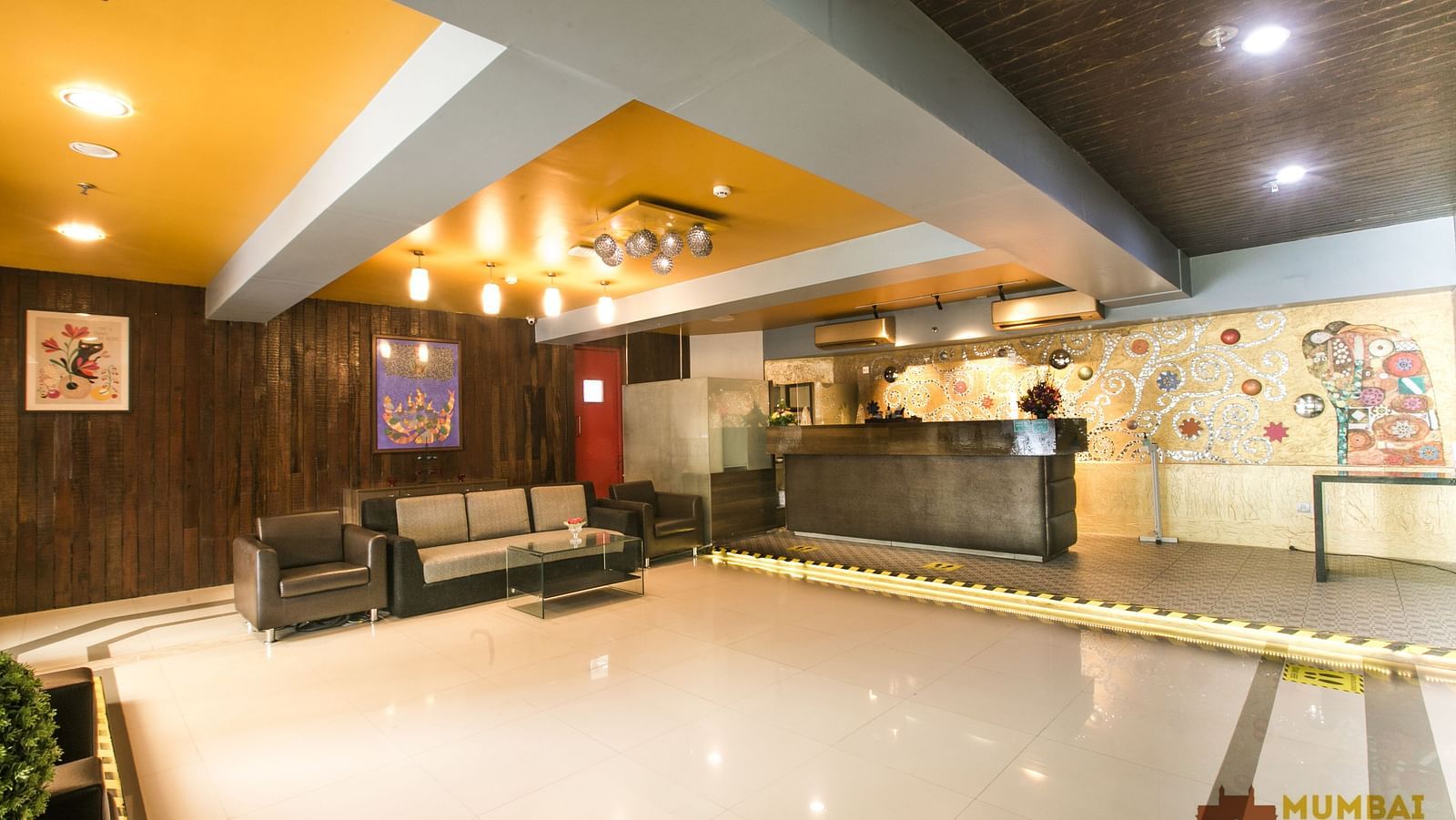 Hotel lobby with colorful wall art, a yellow accent ceiling, seating area, and reception desk with a mosaic backdrop.