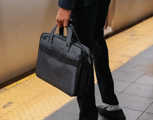 Person in formal wear on a platform by a train, holding a briefcase. Textured tiles and safety marking visible.