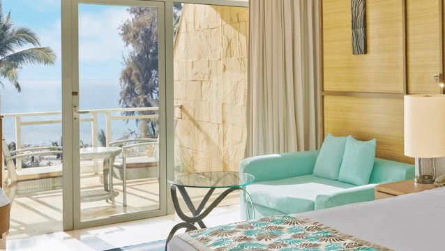 Bright, airy room with a mint-green sofa, glass coffee table, and a balcony offering a clear view of the sea and palm trees at The Resort, Mumbai, the perfect place for a Couple Staycation near Mumbai