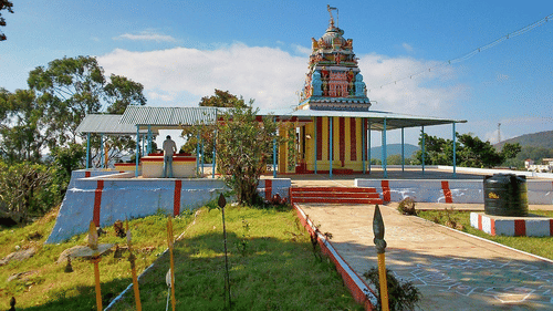 A colourful Hindu temple with a detailed tower stands under a bright blue sky. A person is standing near the entrance. The temple is surrounded by greenery, hills, and a walkway leading to the structure.