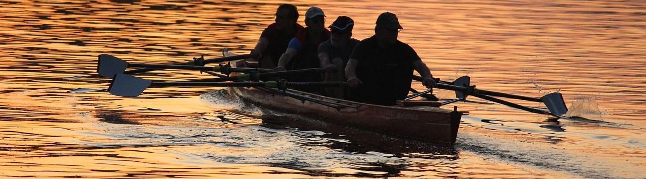 People rowing a boat with oars over a lake reflecting a golden sunset.