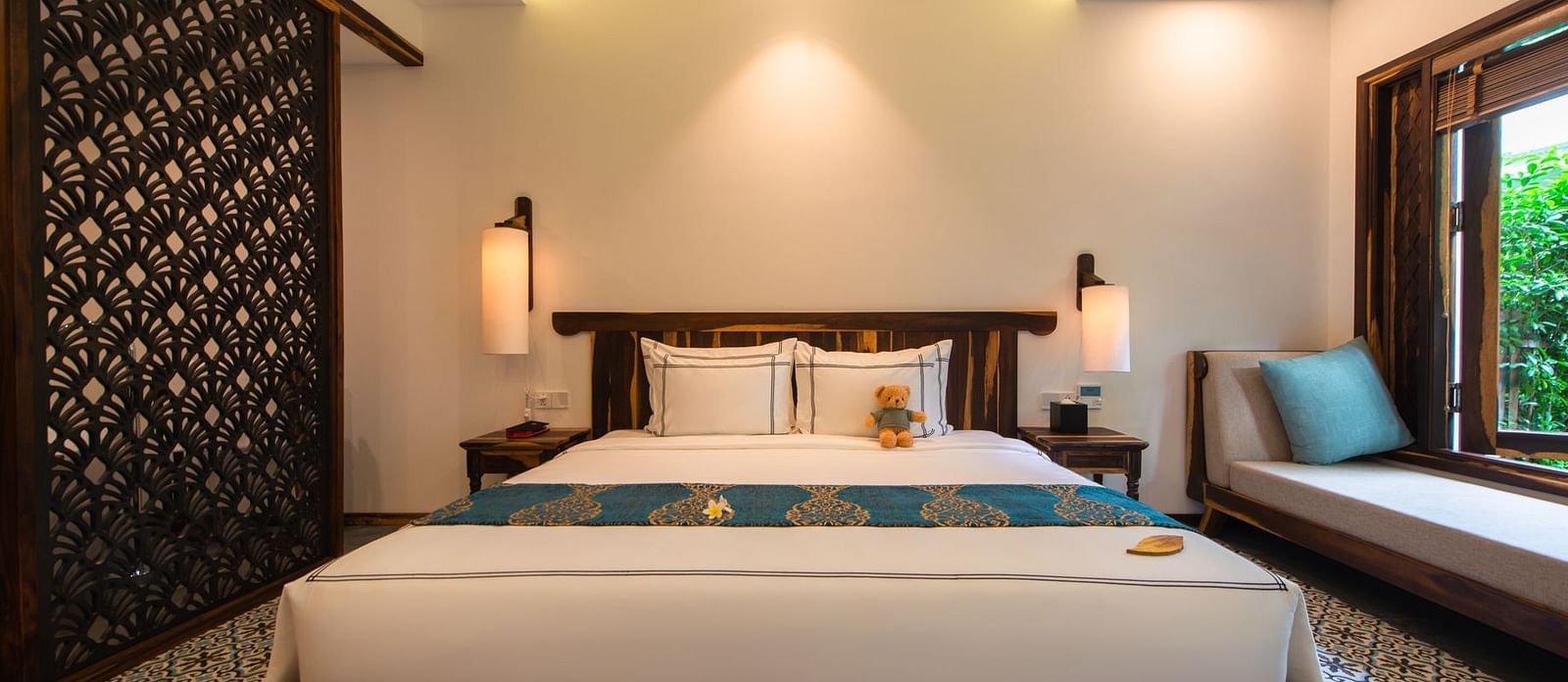A front view of a large white bed with a teddy bear on it with a balcony to the right - Alibu Resort Nha Trang
