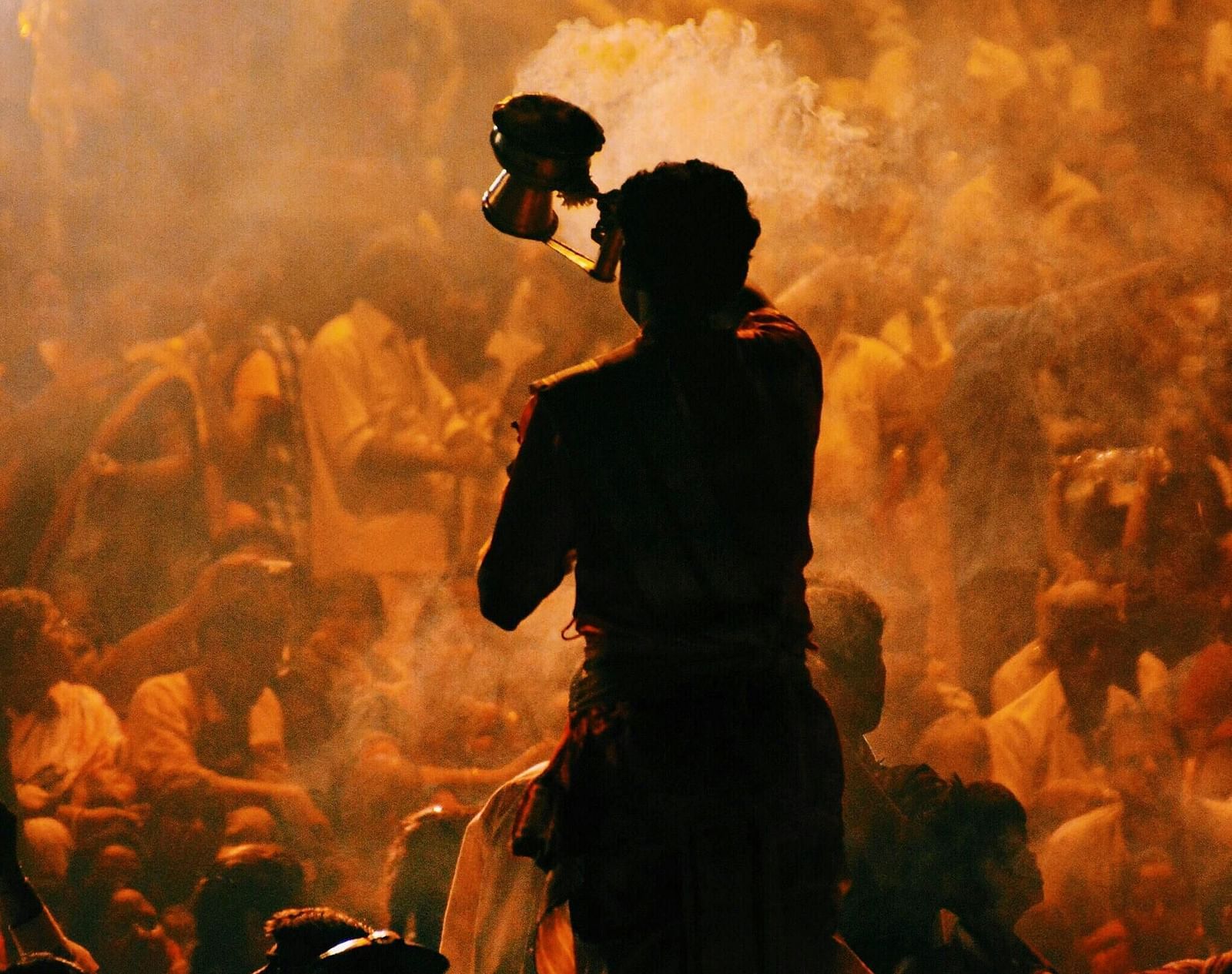 A silhouette of a priest performing aarti with several devotees in the background.
