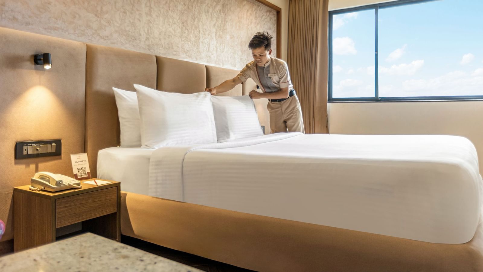 A person arranging pillows on a large bed in a room at Mango Hill Central, Chennai. There is a large window on the right.
