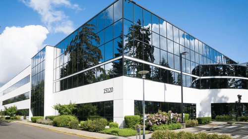 A modern office building with reflective glass windows and white panels stands under a bright blue sky with clouds.