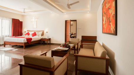 Bright hotel room with a well-made bed featuring red accents, a comfortable seating arrangement with armchairs and a sofa, shiny, reflective tiled floors, and a view into another area - Jenneys Residency, Coimbatore
