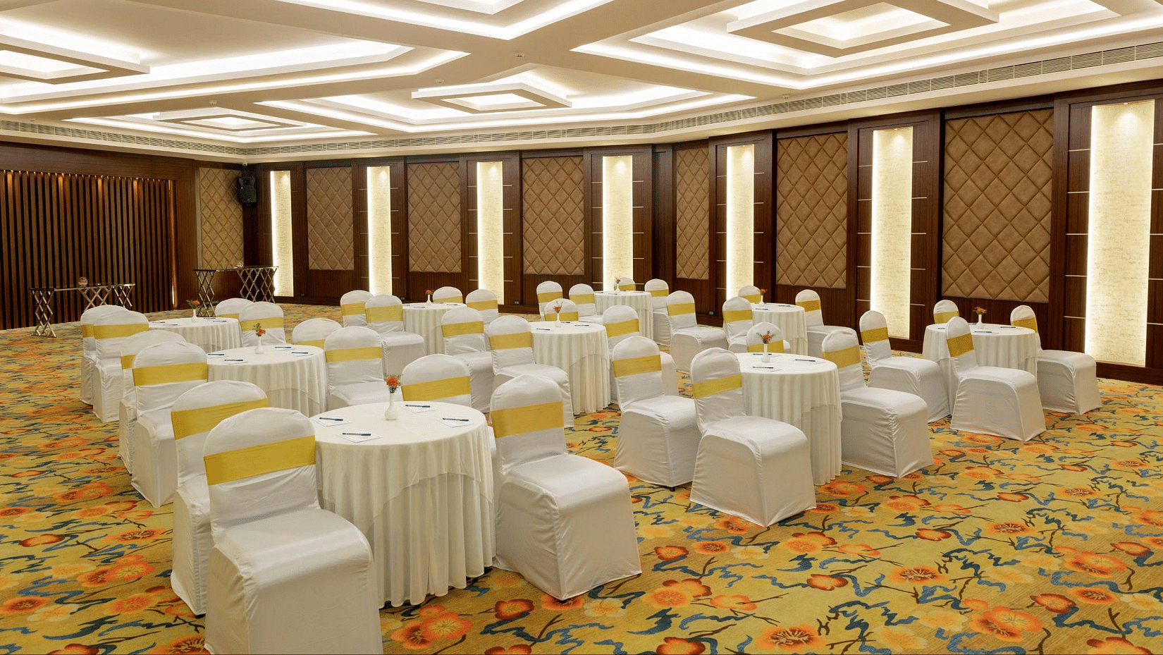 Banquet hall with round tables draped in white, chairs with yellow sashes, patterned carpet, and a geometrically designed ceiling - Jenneys Residency, Coimbatore