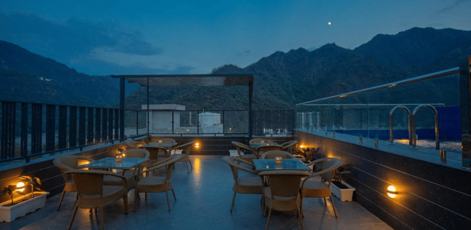 A dimly lit outdoor rooftop restaurant area with several tables and chairs, and a pergola, set against a backdrop of mountains under a twilight sky at Perfectstayz Premium Laxmi Heritage.