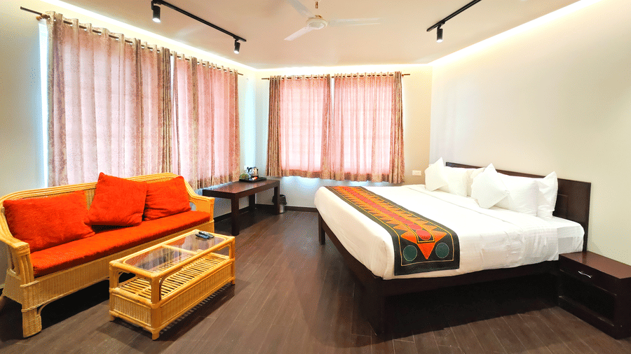 Bedroom in Pipit room at Ibex Resorts, Coonoor (Tapas). The room features a bed positioned next to a bedside table, a sofa with a coffee table, and windows covered by curtains.