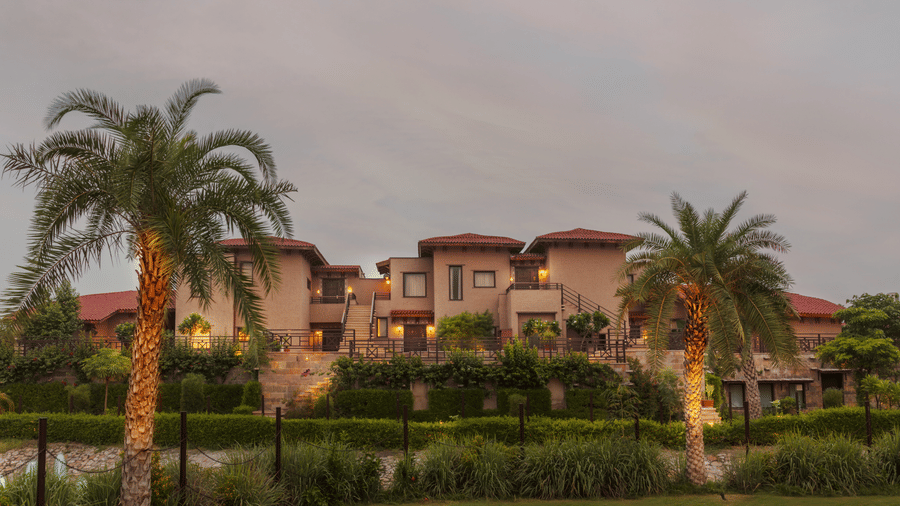 The resort's facade as seen from a distance at dusk, with lush greenery and palm trees under a cloudy sky at Aamaghati Wildlife Resort. Get in touch with Ranthambore Resorts Contact mentioning on the contact us page.