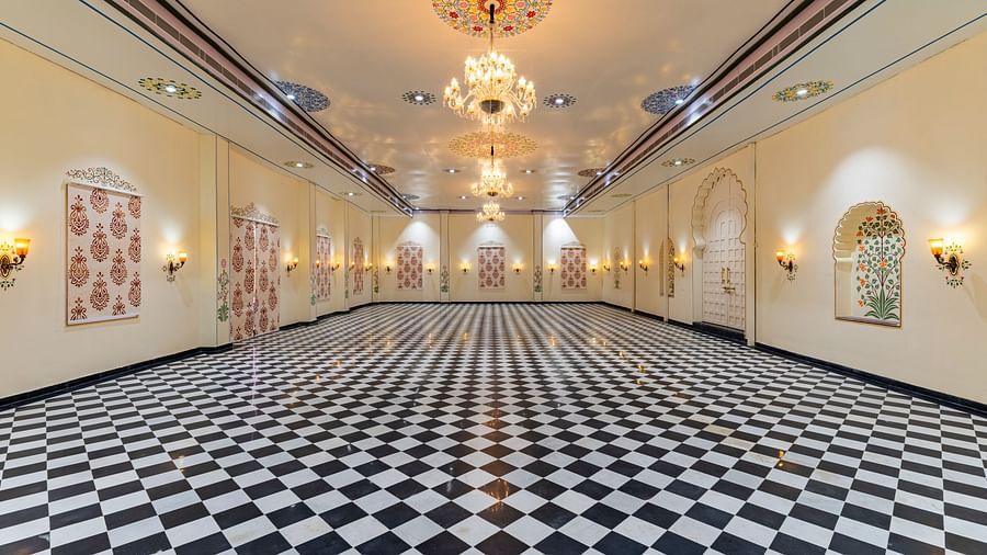 Long hallway with black-and-white checkered flooring, ornate golden chandeliers, and framed artwork on cream walls.