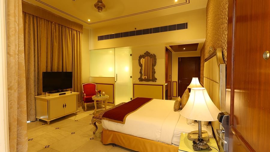 Image of a room with a bed, a television in front and lamps on the side of the bed at Rajasthali Resort & Spa Jaipur.
