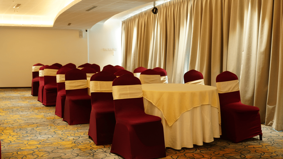 A conference setup featuring a round table with chairs covered in fabric alongside a long curtained window at Tiara by MPS.