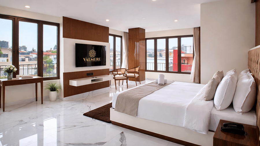 A spacious Junior Suite AC Room at Valmere Chery Blossom Suites featuring a bed with multiple pillows, a flat-screen television facing the bed, and large windows offering views outside.