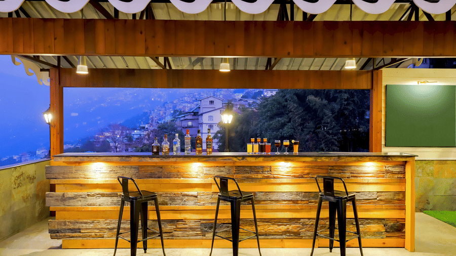 Bar at Valmere Himalayan Hotel & Spa, Gangtok with a wooden counter, bottles of spirits, 4 colourful cocktails, 3 stools, and a hillside town view at dusk.