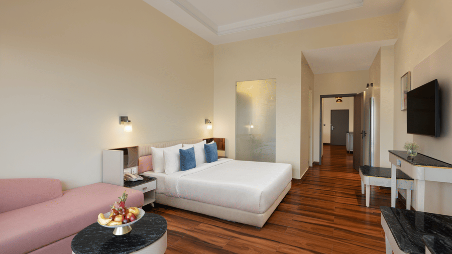 A room with a large bed, a sofa, a table spreads, pillows, a television, and windows at Essentia Luxury Hotel and Convention, one of the best hotel in Nagpur.