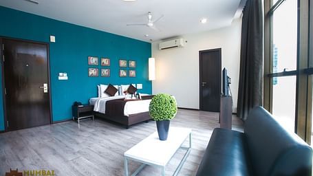 Spacious hotel room with a seating area, wooden floor, turquoise accent wall with art, a white coffee table, and a window.
