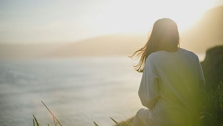 A woman sitting on a cliff while watching the sun near a beach in the distance.