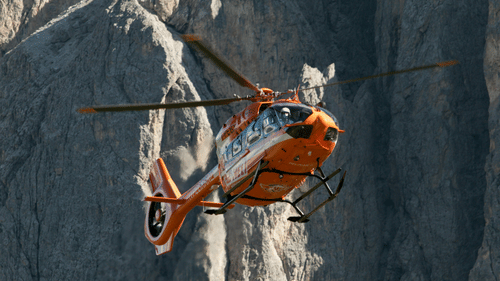 An orange and white helicopter flying close to a steep, rocky mountain face under bright daylight at Ananta Express, Katra.