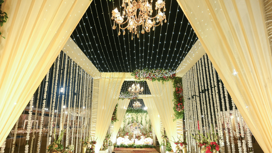A ceremonial aisle setup with hanging string decor, draped fabric panels, chandeliers above, and a stage at the far end.