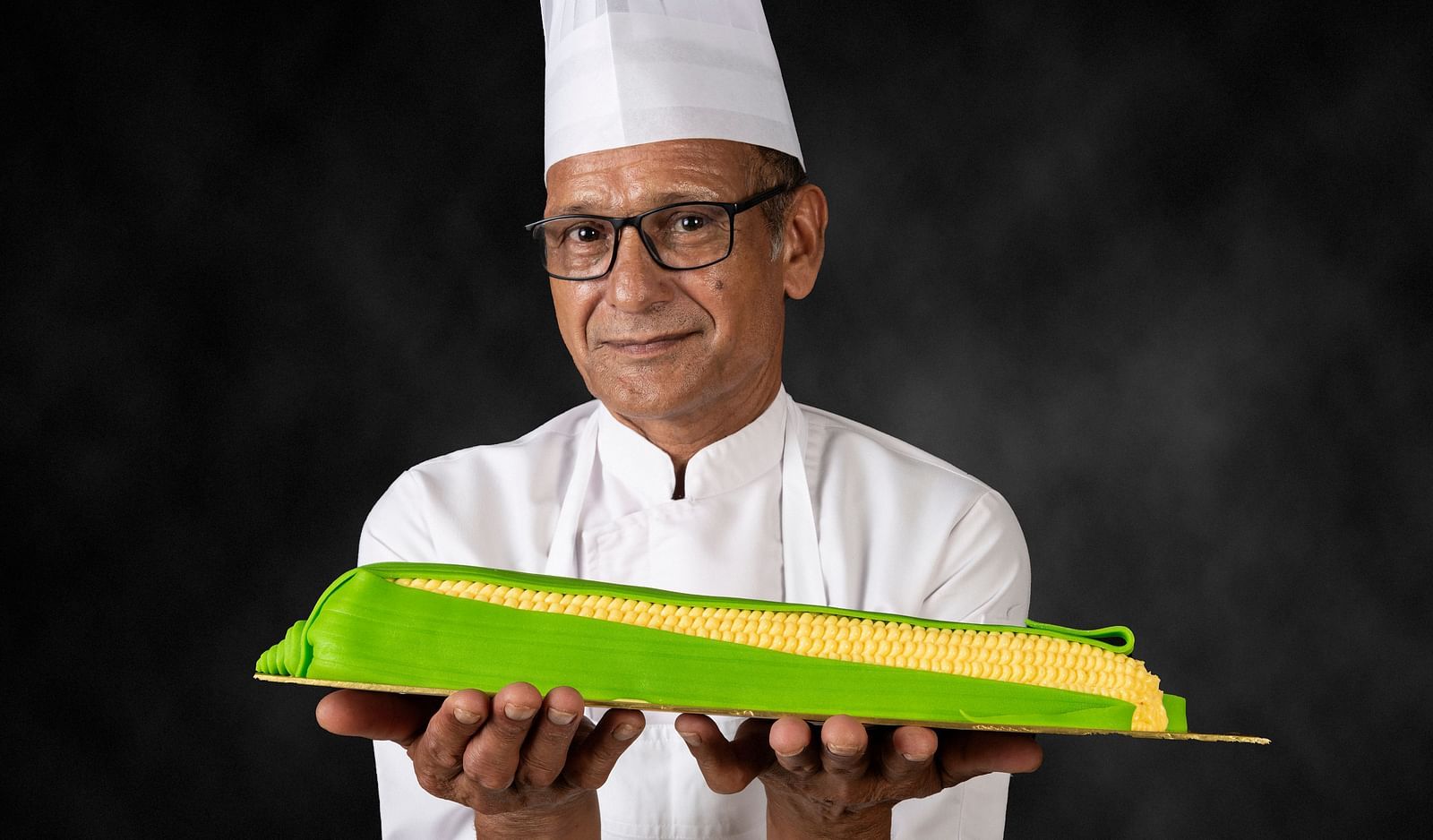 The image shows a chef wearing a white uniform and a chef's hat, holding a creatively designed corn-shaped dessert on a plate