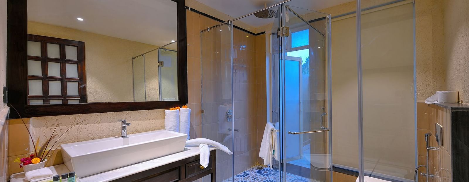 A modern bathroom of Premium Suite Cottage with a rectangular sink, a large mirror, a glass-enclosed shower with patterned tiles, a western toilet, and rolled white towels - Singhvilas, Ranthambore