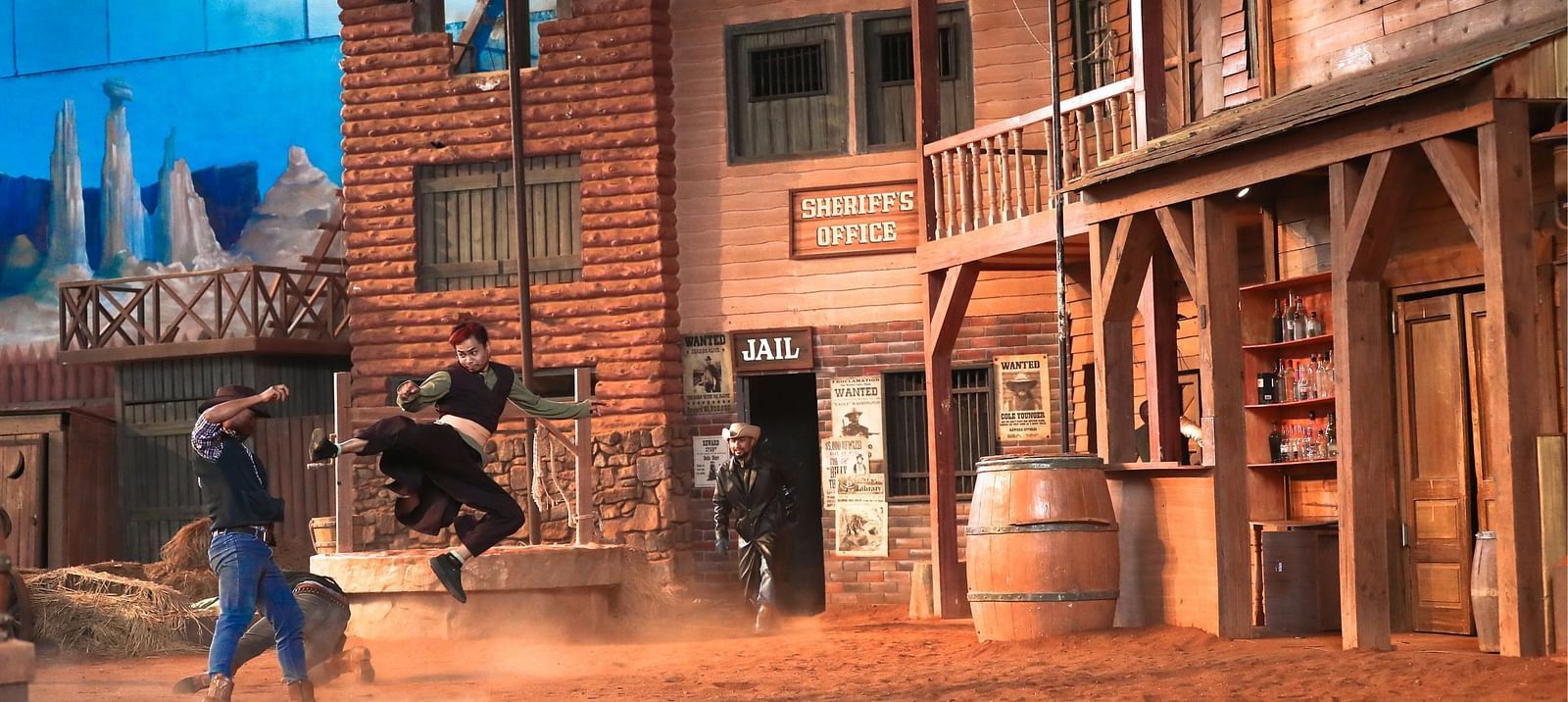 Wild West film set scene with actors in action. One in cowboy attire swings on a rope, while another runs beneath. Background features rustic buildings, wooden barrels, and a dusty ground. Sky backdrop visible above the set.