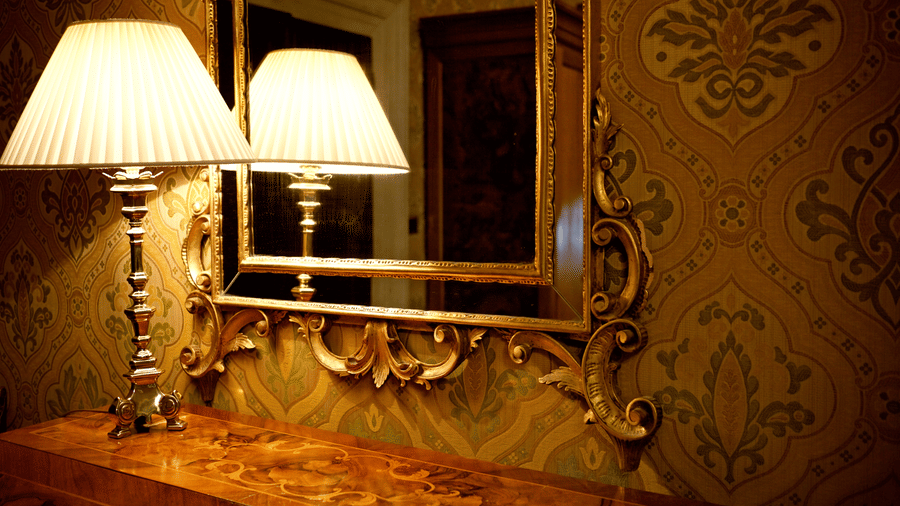 A patterned wall with a desk in front, a warmly lit table lamp, and a mirror on the wall