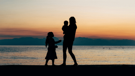 The silhouette of a mother and 2 children is framed against a soft sunset glow on a peaceful beach.