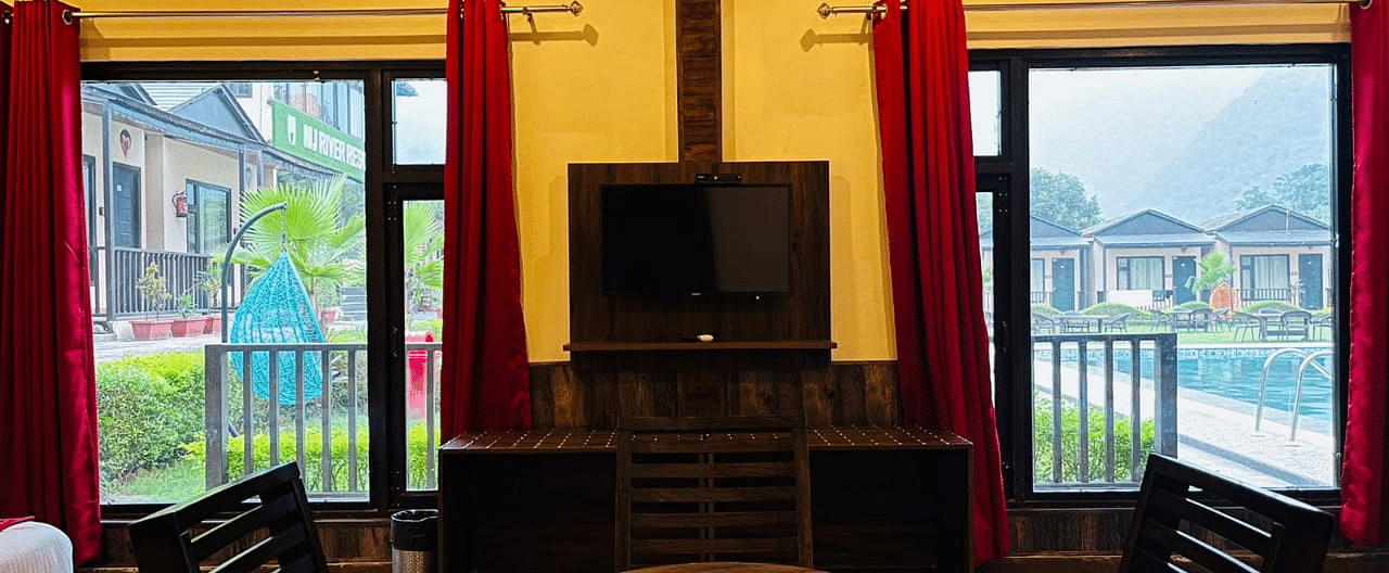 View of the dining table placed in front of a TV with outdoor views on either sides in Family Cottage at DLS MJ River Resort, Rishikesh.