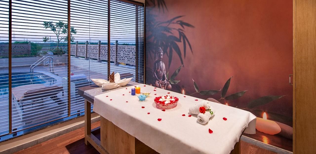 A serene spa treatment room featuring a massage table draped with a white sheet and adorned with red rose petals and decorative items, set against a warm-toned wall with a subtle mural and with natural light coming through large windows overlooking an outd