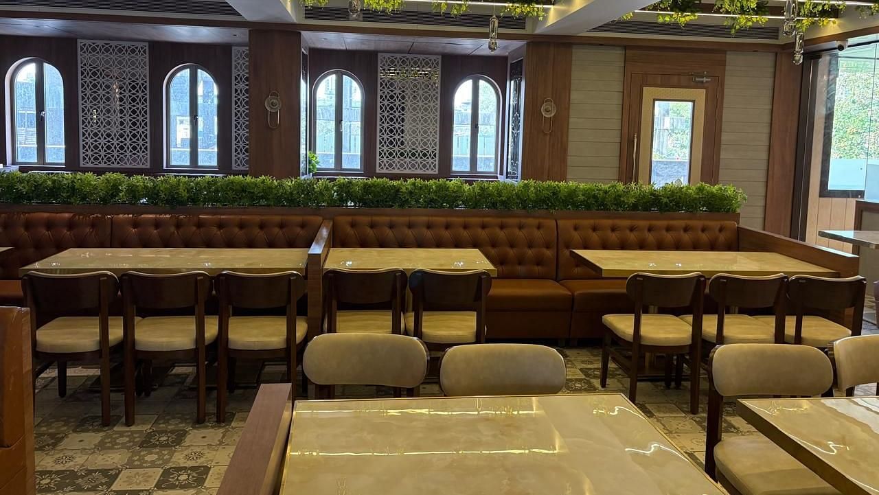 A brightly lit restaurant area with wooden tables and chairs, featuring arched windows and some greenery - Melior Inn