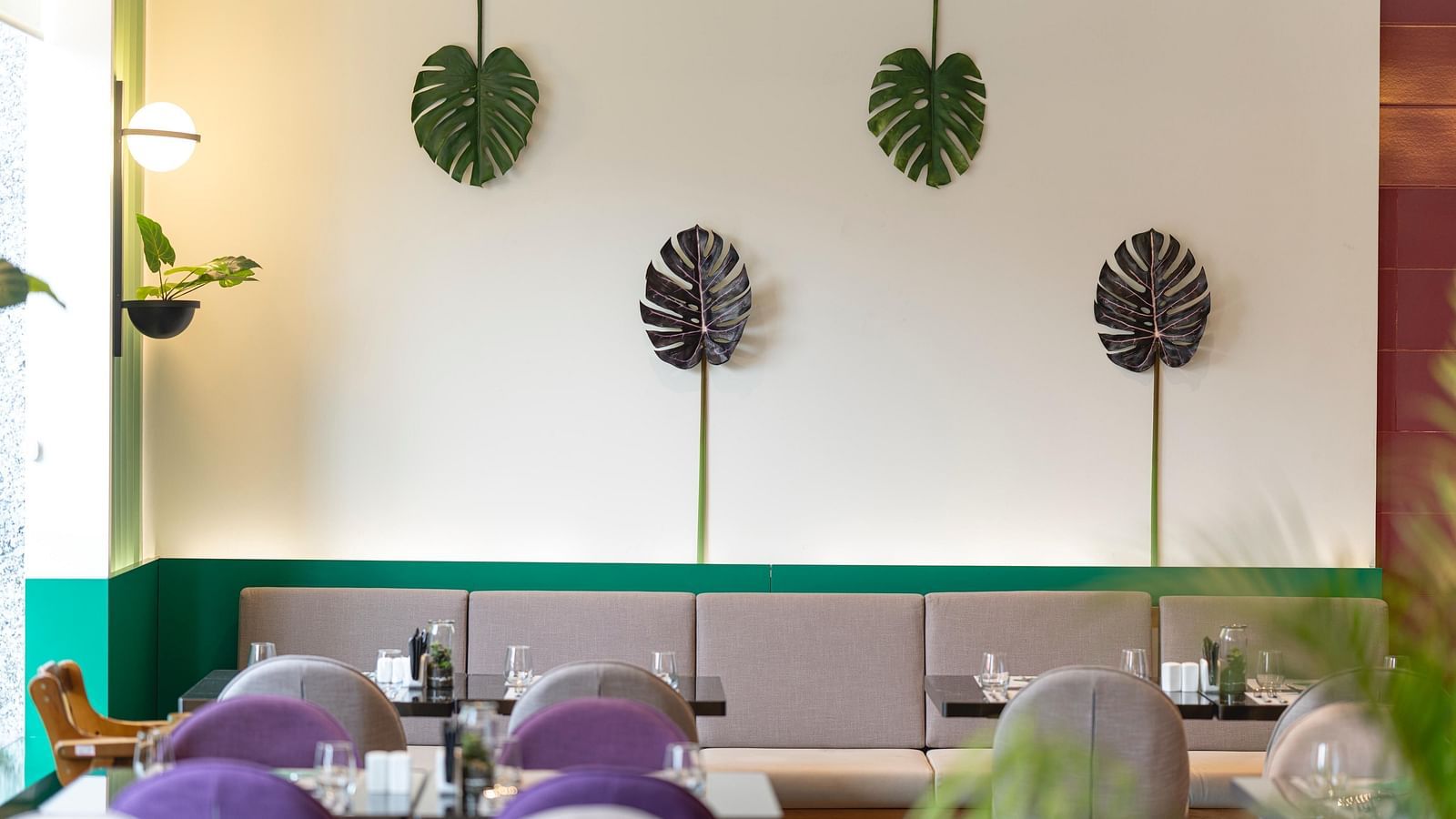Bright, modern Meli-Melo restaurant interior with accents, large windows, potted plants, and neatly set tables with chairs and beige booth seating. Natural light fills the stylish dining area