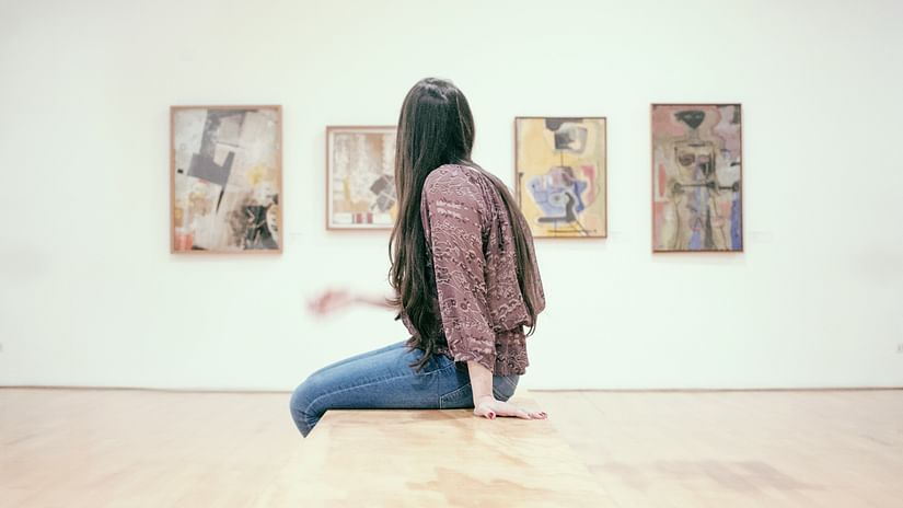 A woman sitting on a bench and looking at the paintings hung on the wall in the distance