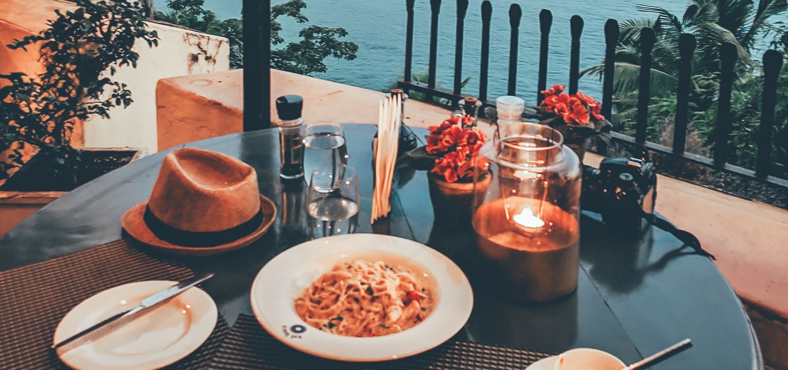 An outdoor dining table on a balcony at Fort Tiracol, Goa, set with food and drinks, overlooking a scenic view of a bay and distant land under a light sky.