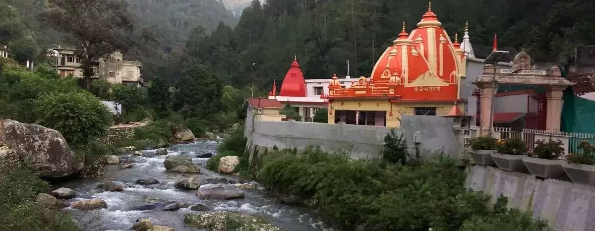 A temple situated on the banks of a river which has small boulders and rocks visible, pictured with the backdrop of the majestic mountains.