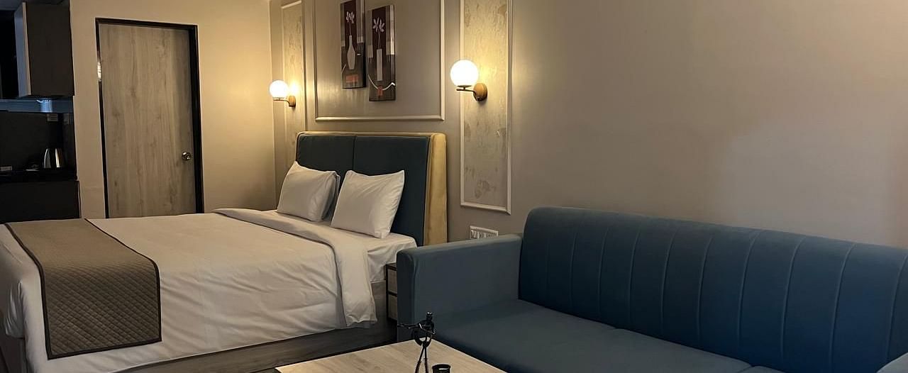 A spacious room featuring a large bed with neatly arranged pillows and lampstands, a cushioned sofa, and a table placed in front of it at Urban Suites by BluSalzz, Jaipur.