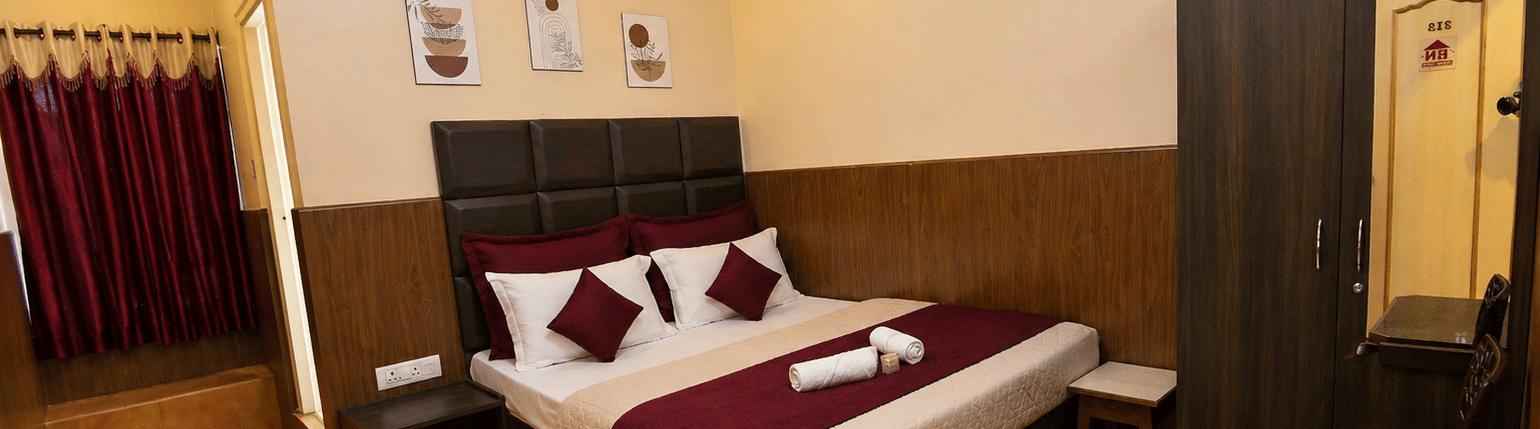 A neatly-made bed with towels, a tall wooden wardrobe, and a small bedside table in the Deluxe Double Room With Extra Cot at BN Tourist Home, Ooty.