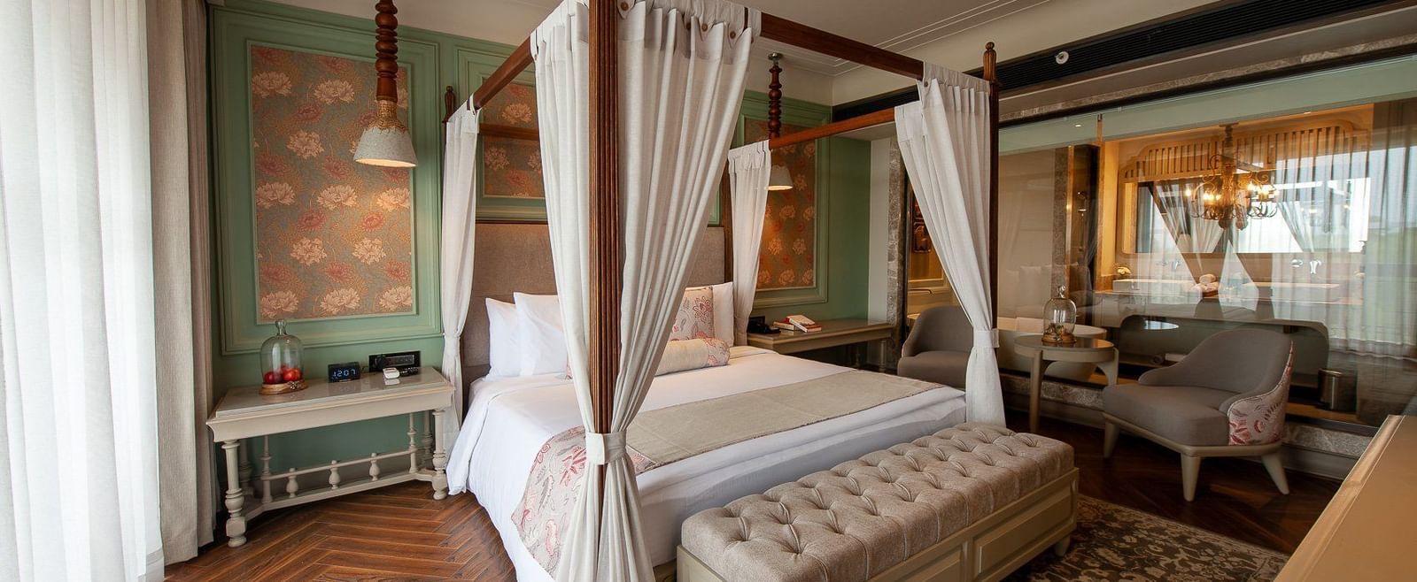 A wide view of the elegant suite at The Evren, Vagator, showcasing a four-poster bed, a stylish rug on a herringbone floor, and a seating area with two armchairs near a window. The room has a view into the connected bathroom.