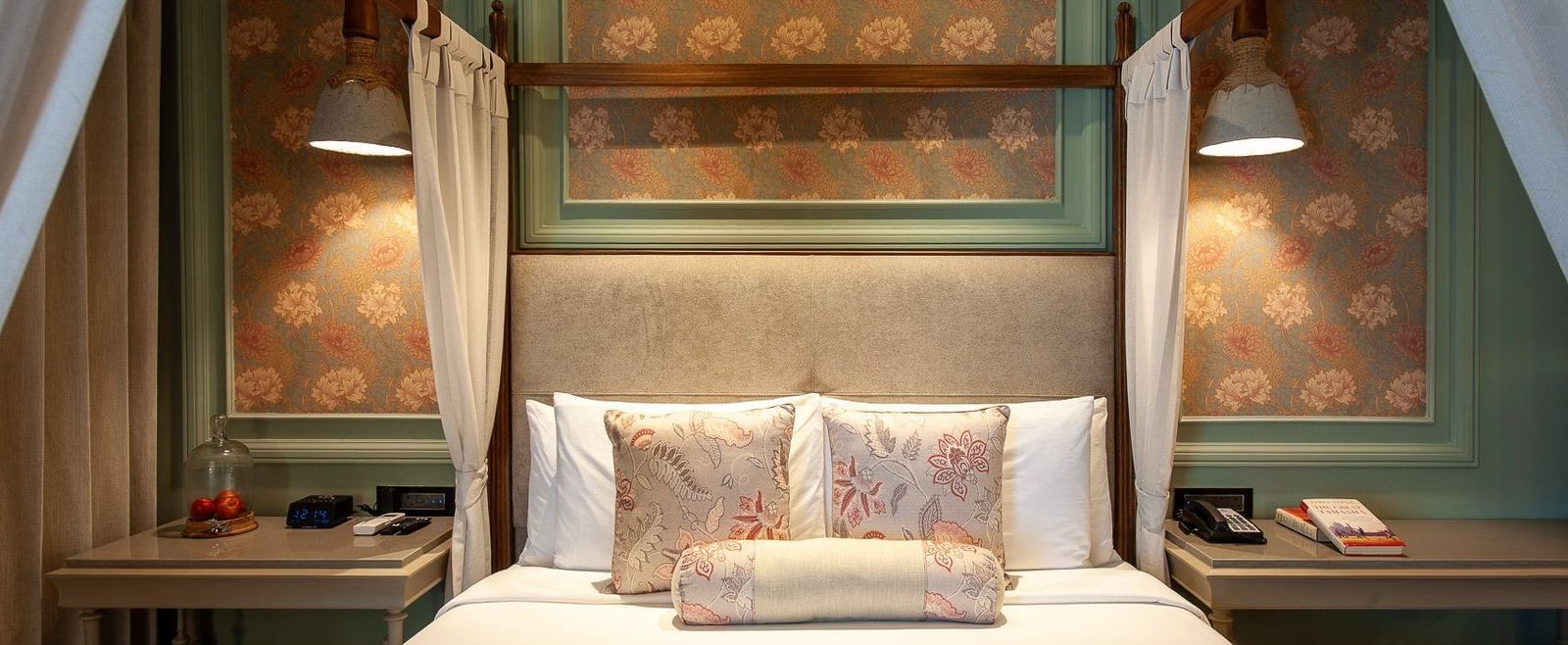 A close-up, front-facing view of the beautifully appointed four-poster bed in a suite at The Evren, Vagator. The bed is adorned with decorative pillows, and the background features elegant wallpaper and unique hanging lights.